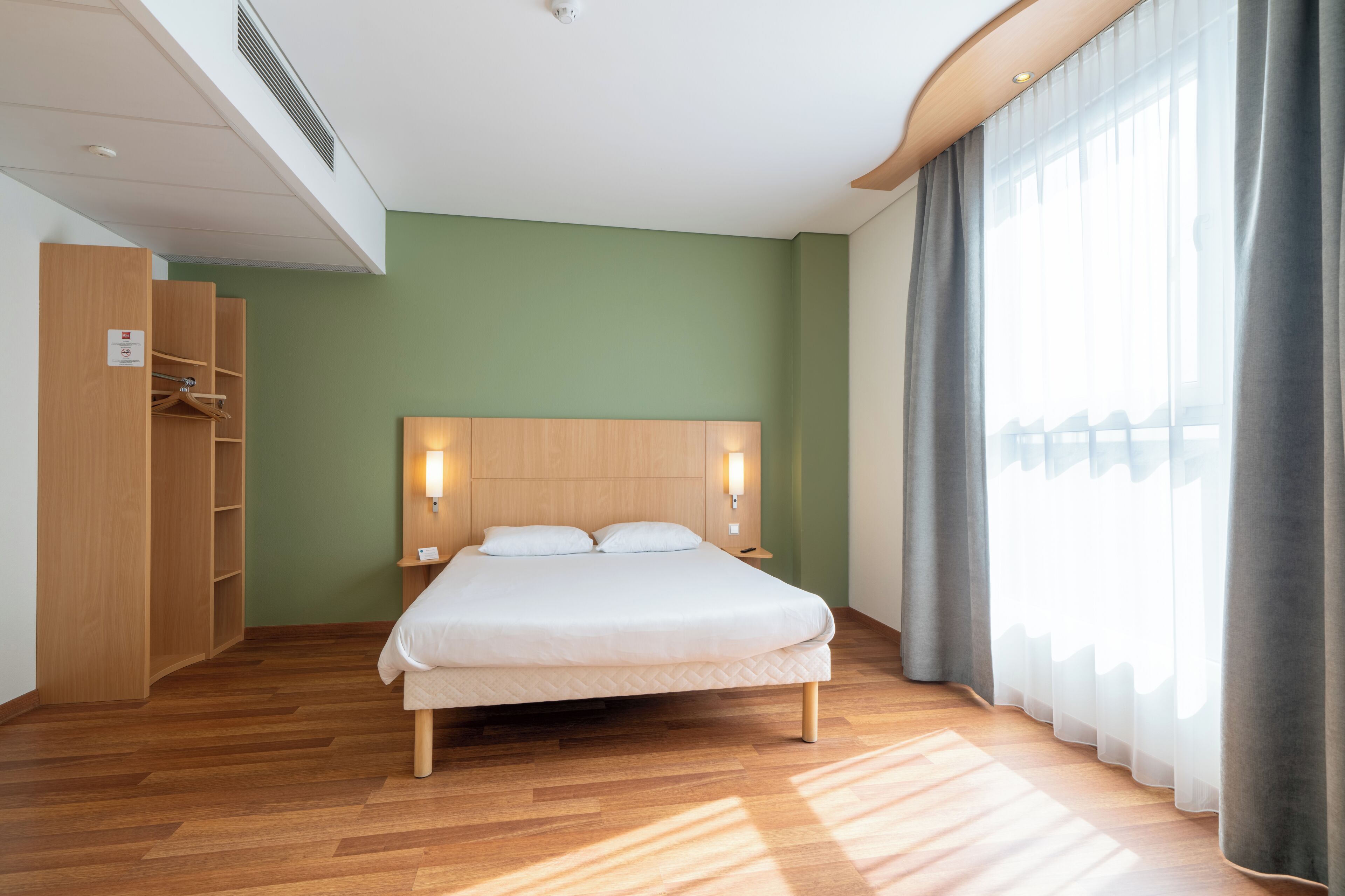 Una habitación de hotel moderna con una cama de madera, un armario y un ventanal con cortinas.