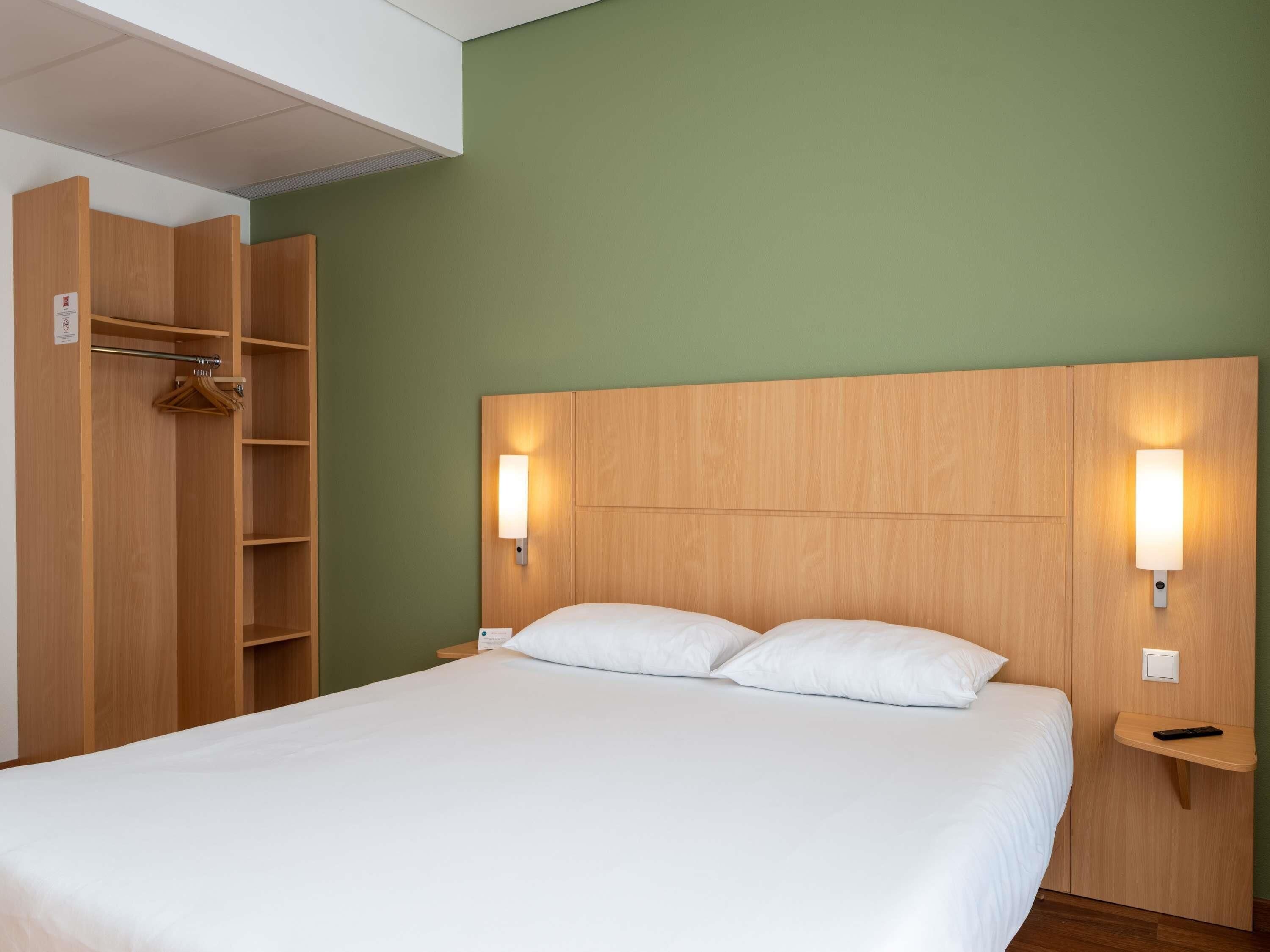 A hotel room with a bed, wooden wardrobe, and two wall-mounted nightstands.