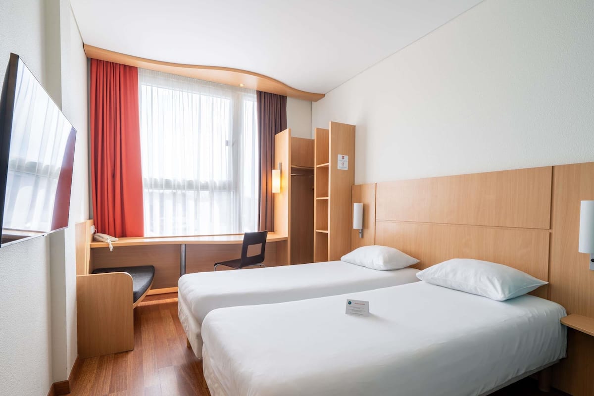 A hotel room with two beds, a desk, a chair, a TV, and a window with curtains.