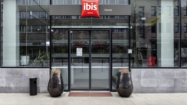 The exterior of an ibis hotel with a modern glass facade, a red logo, and two large vases in front of the entrance. - ibis Kaunas Centre (Kaunas)