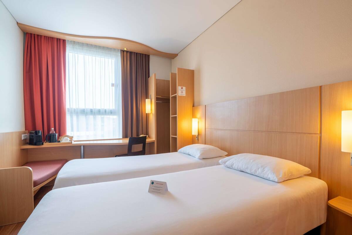 A hotel room with two beds, a desk with a chair, a window with curtains, and a wardrobe.