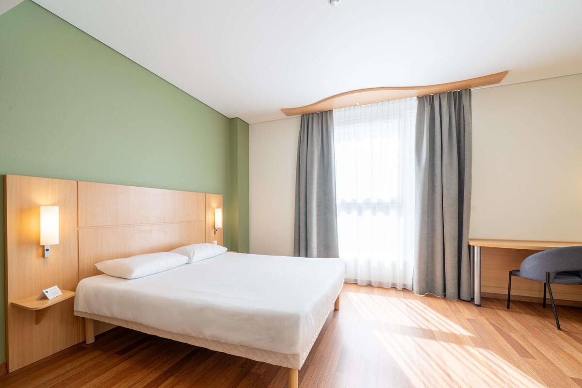 A hotel room with a bed, bedside tables, a desk, and a window with curtains.