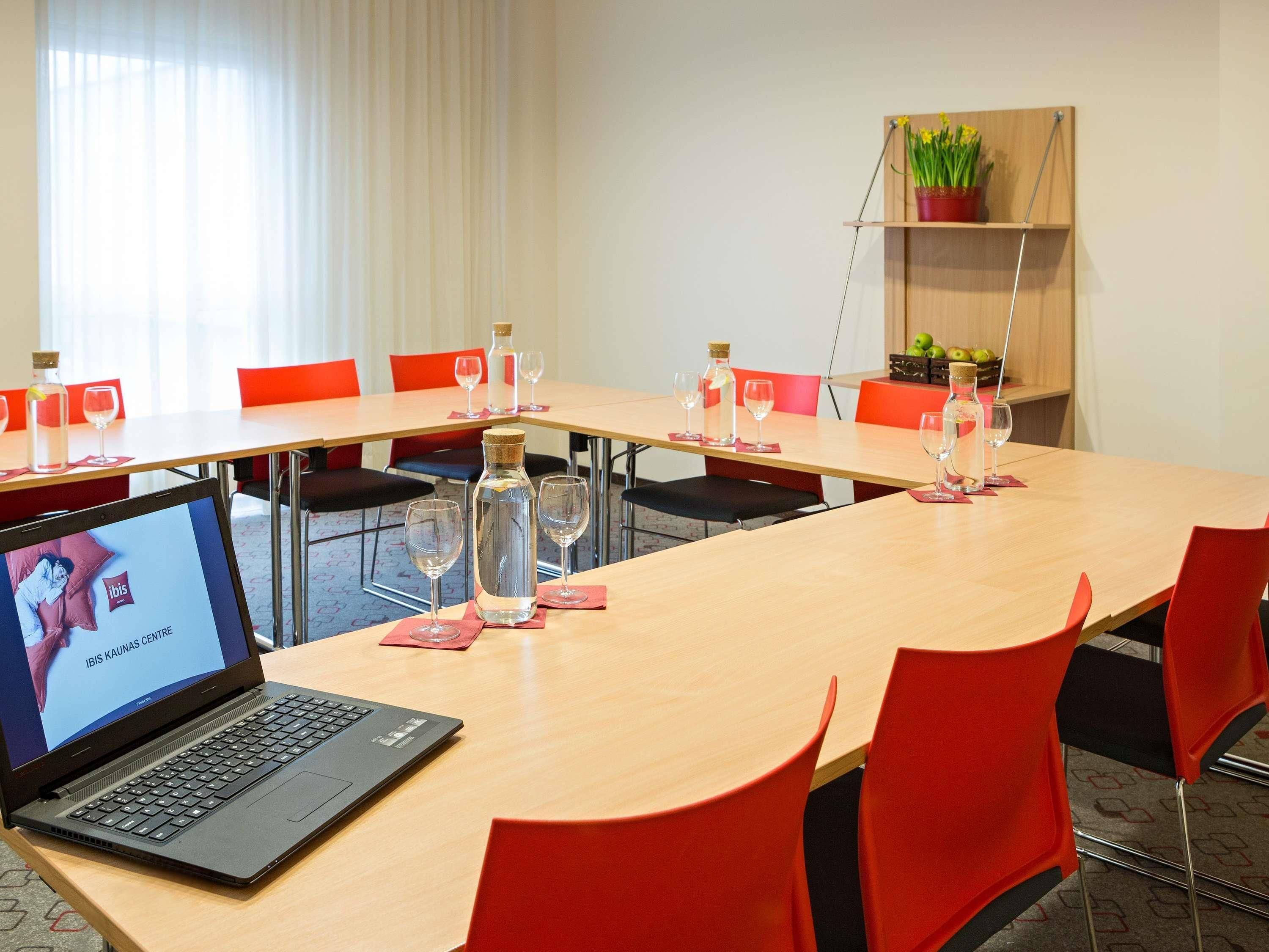 Una sala de conferències amb un ordinador portàtil, cadires vermelles, una taula llarga i una planta en maceta sobre una prestatgeria.