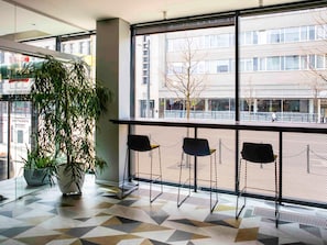 A modern interior with a geometric patterned floor, a large potted plant, and a view of a cityscape through large windows. - ibis Kaunas Centre (Kaunas)