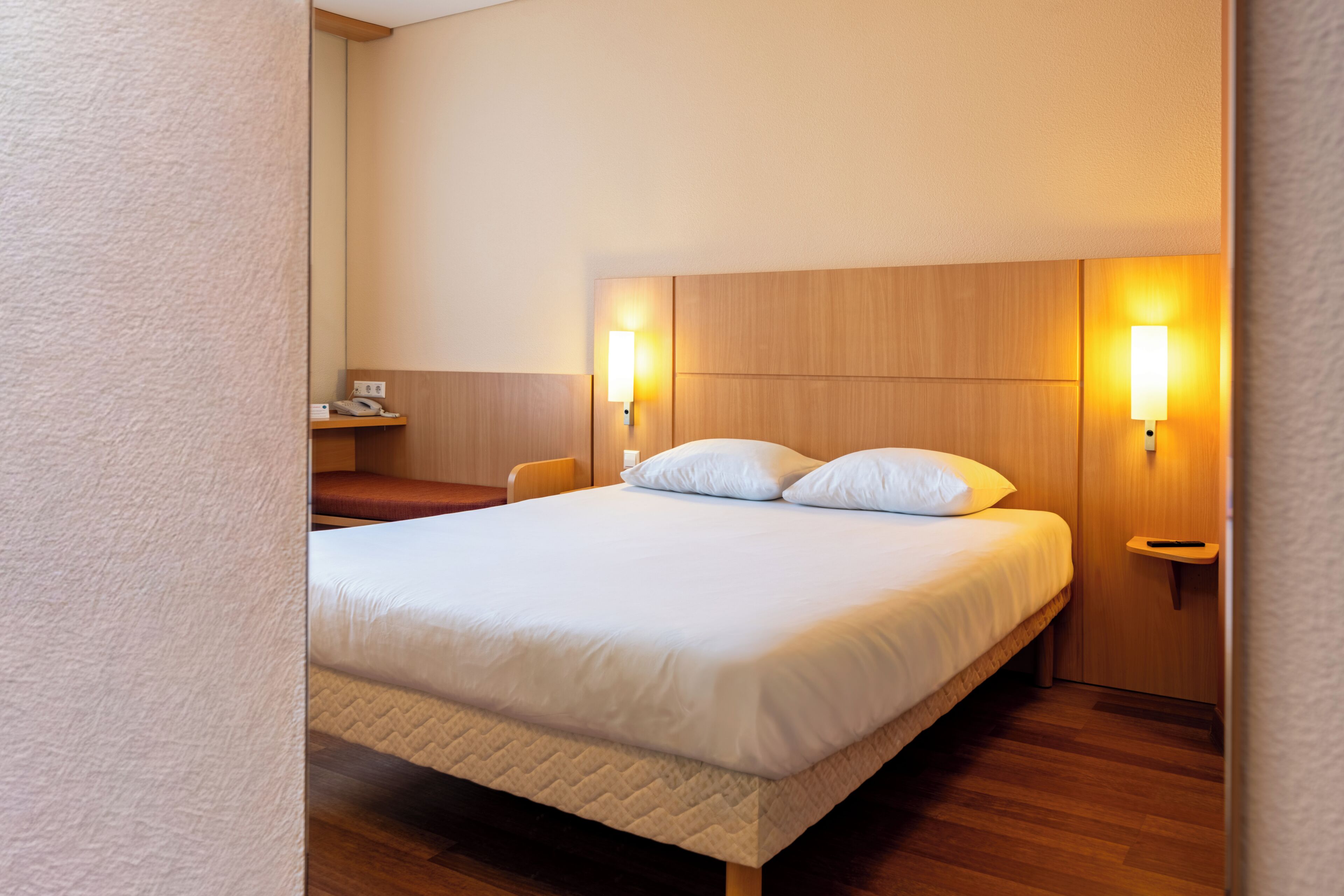 Una cama bien hecha con cabecera, dos mesitas de noche con lámparas y un escritorio pequeño con teléfono.
