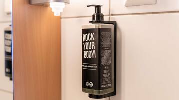 Una botella negra de jabón para manos con dosificador de bomba, etiquetada con 'ROCK YOUR BODY!', fijada a una pared.