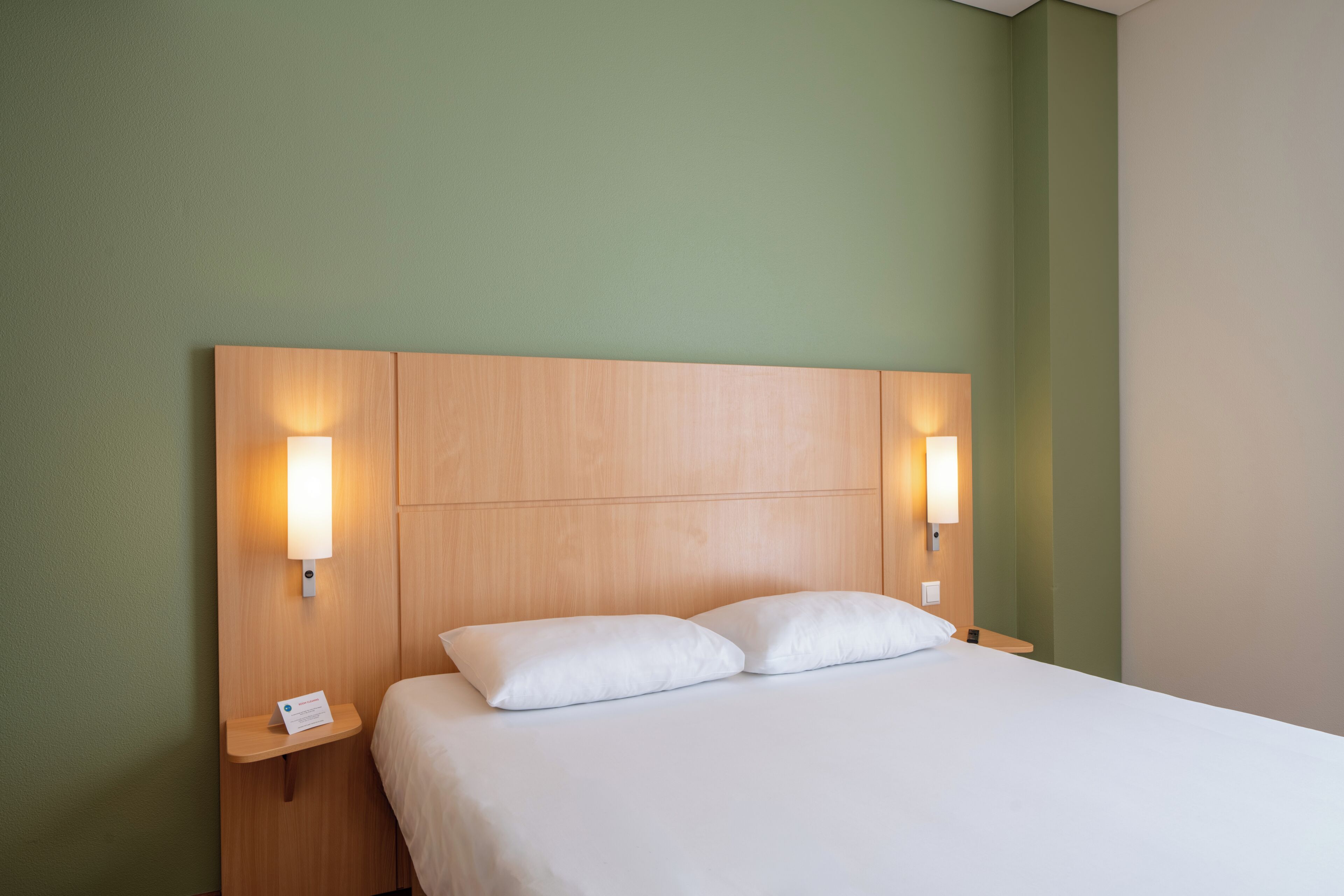 Ein Hotelzimmer mit hölzernem Kopfteil, zwei Nachttischlampen, einem weißen Bett mit zwei Kissen und einem kleinen Nachttisch mit einem Telefon.