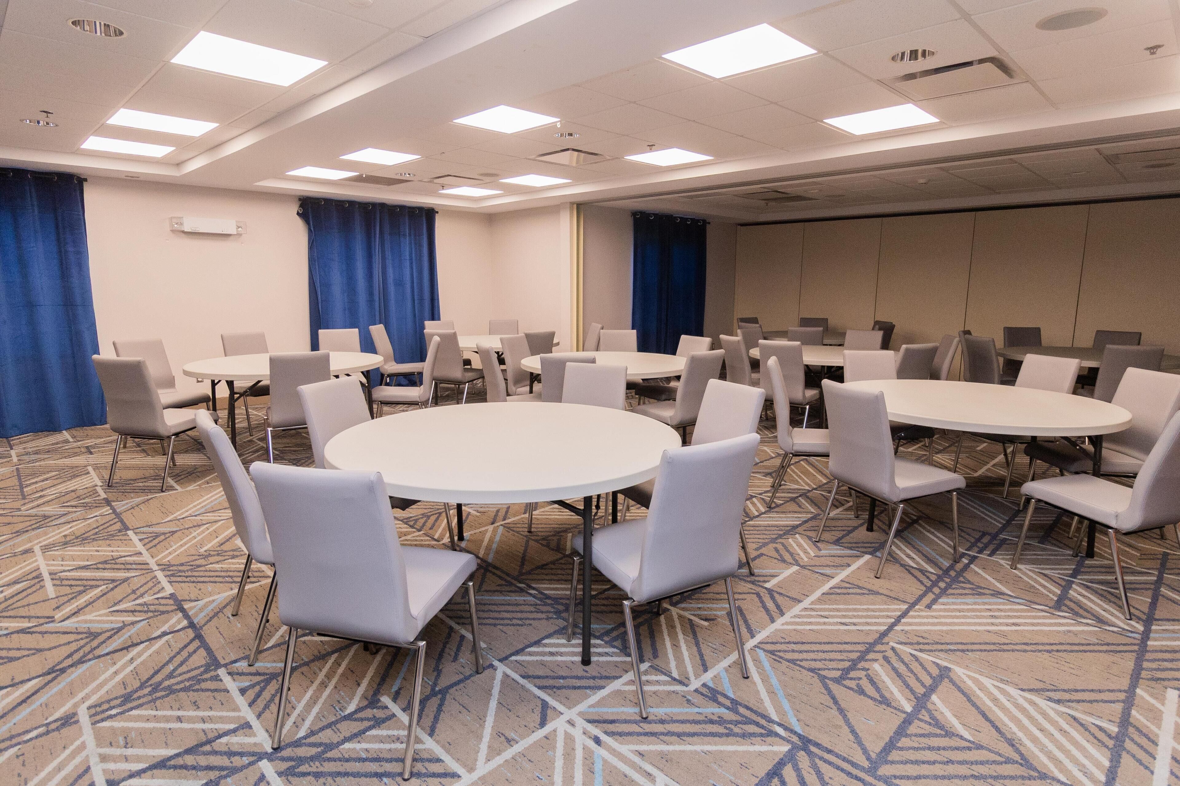 Une salle de conférence dotée de plusieurs tables rondes et de chaises, de rideaux bleus et d’un tapis à motifs.