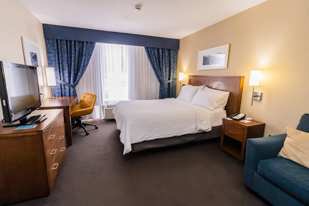 A hotel room with a bed, a desk with a computer, a chair, a TV, and a bedside table with a lamp.