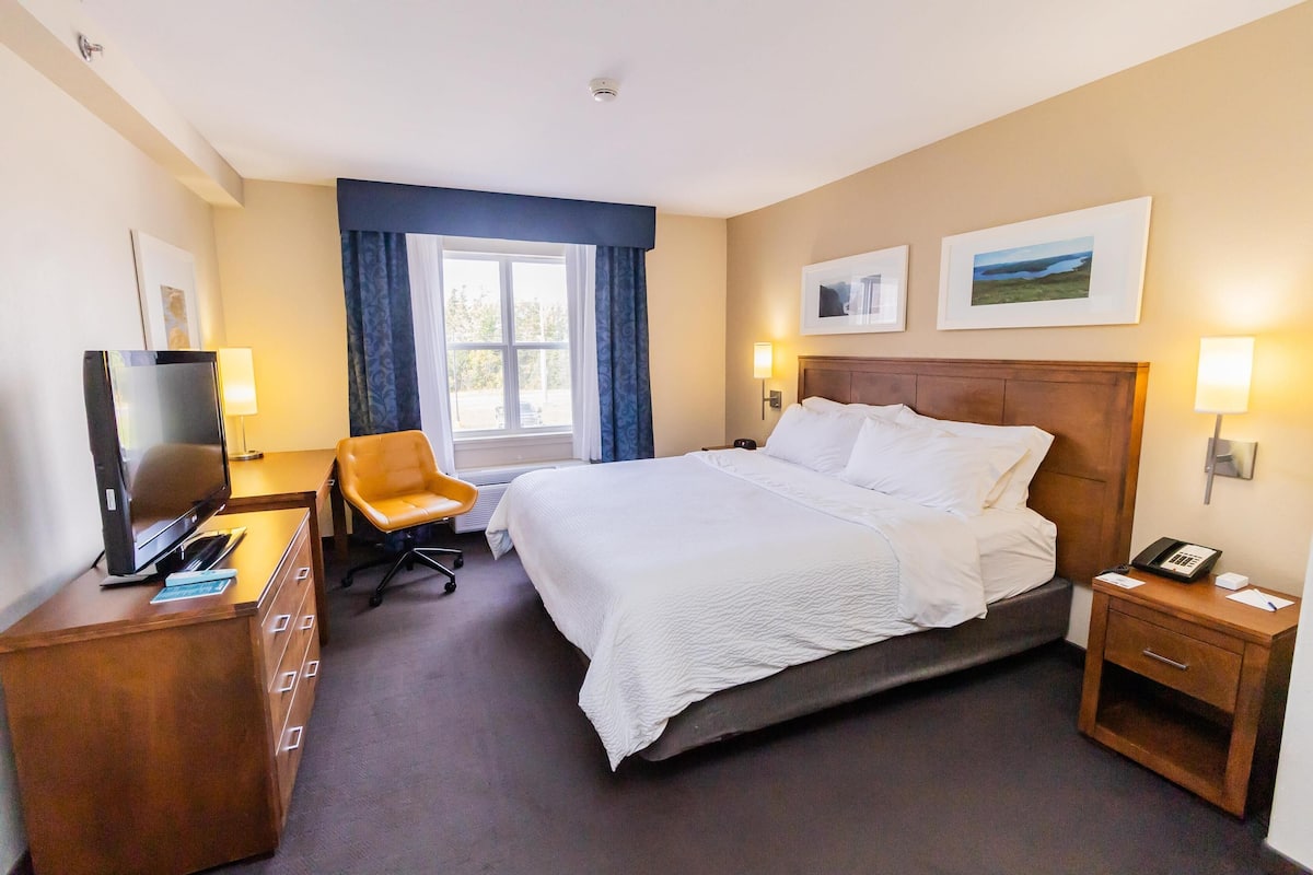 A hotel room with a bed, a desk with a TV, a chair, a nightstand with a phone, and a window with curtains.