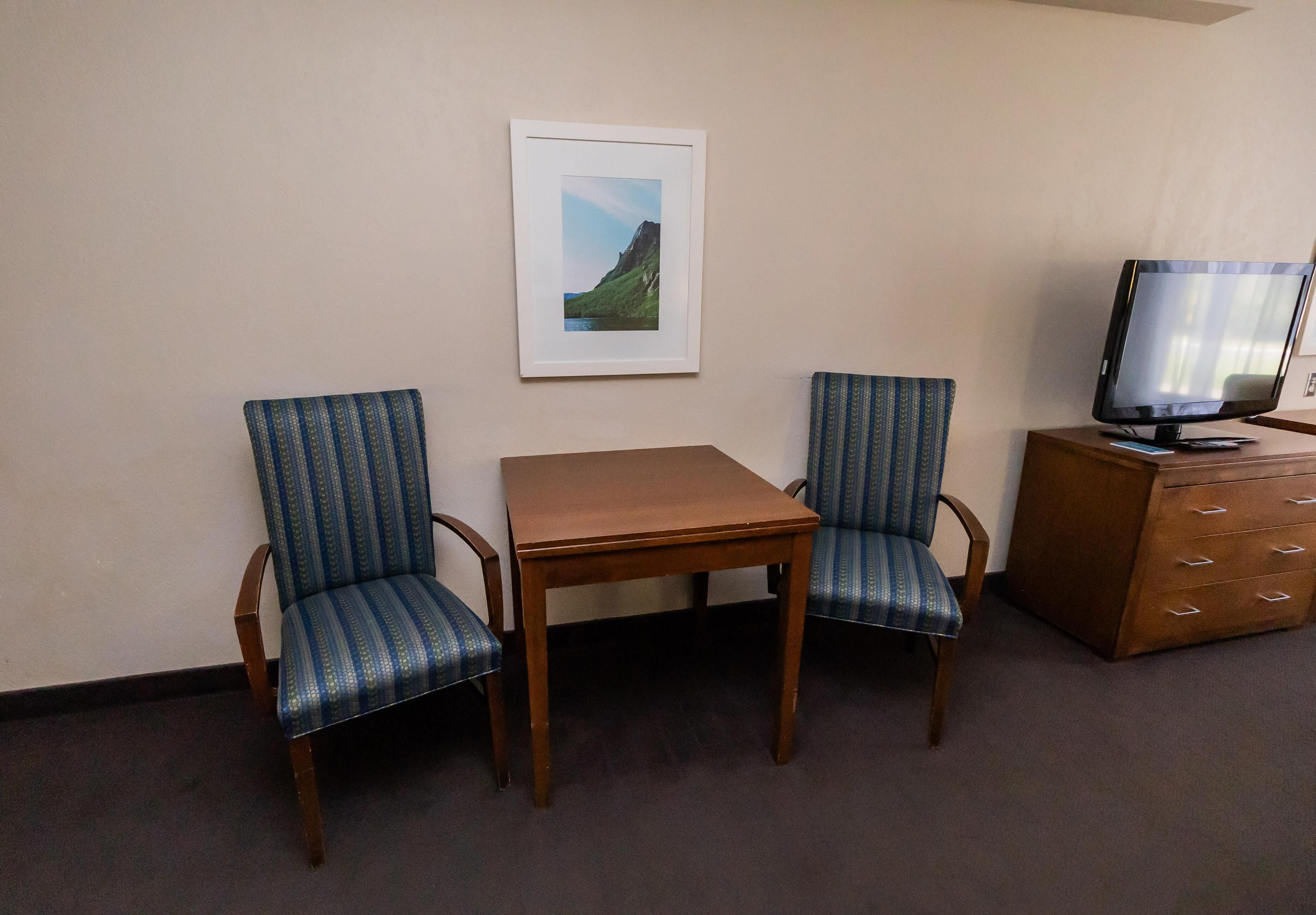 Habitación de hotel con una mesa de madera, dos sillones tapizados, un cuadro en la pared y un televisor sobre un mueble de madera.