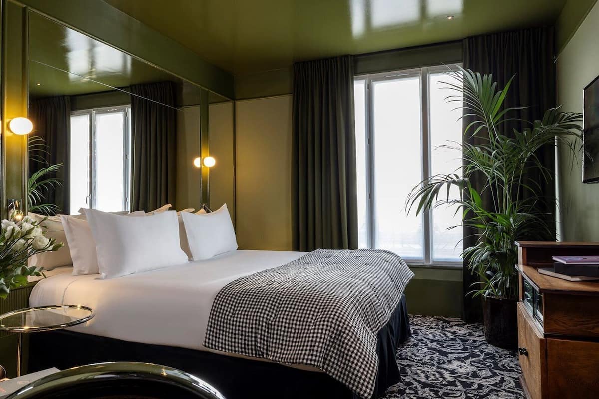 A hotel room with a large bed, a mirror, a bedside table, a dresser, a potted plant, and a window with curtains.