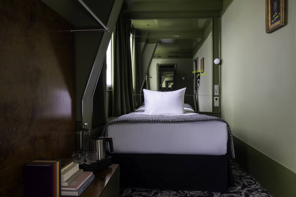 A hotel room with a bed, a bedside table with books and a kettle, a mirror, and a view of the corridor.