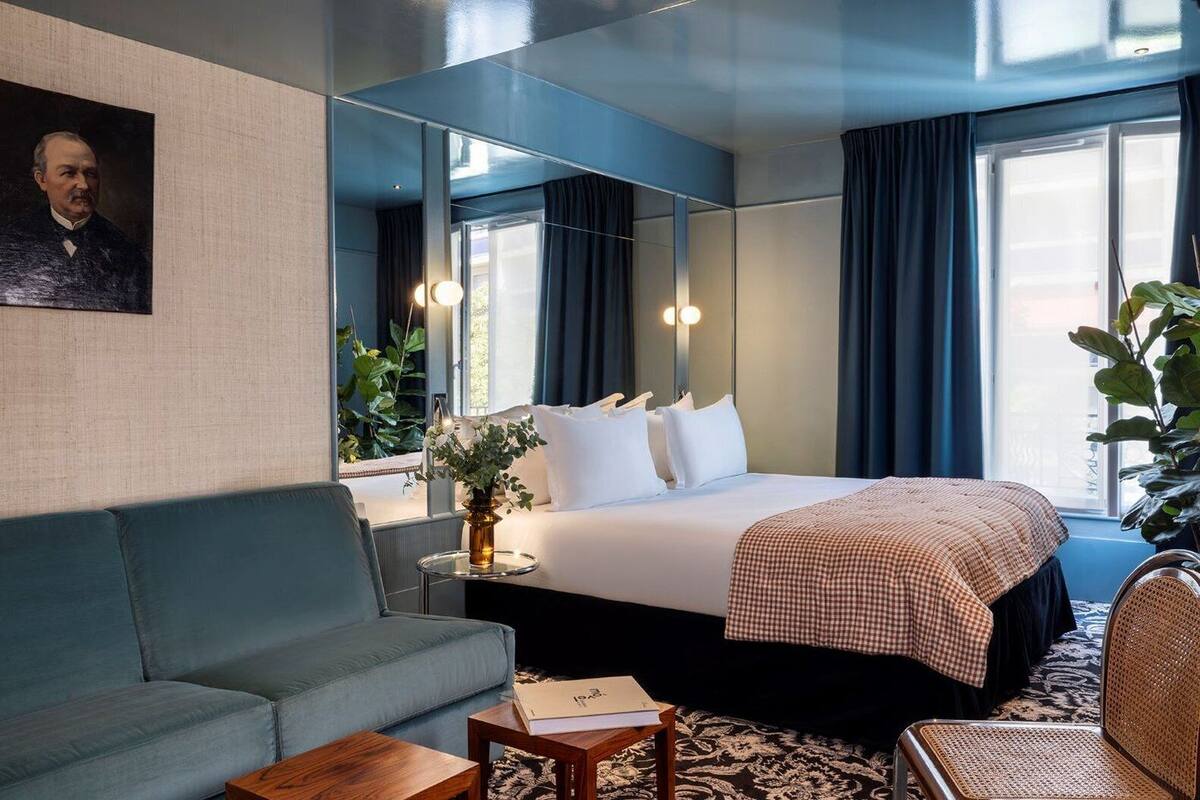A hotel room with a bed, a sofa, a small table, a mirror, and a large window with curtains.