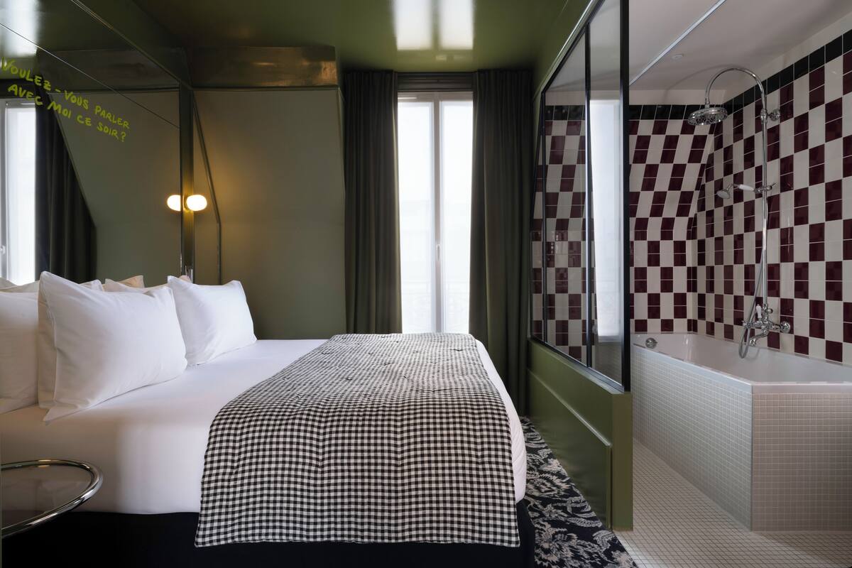A hotel room with a bed, a checkered bedspread, a large mirror, a shower with checkered tiles, and a window with curtains.