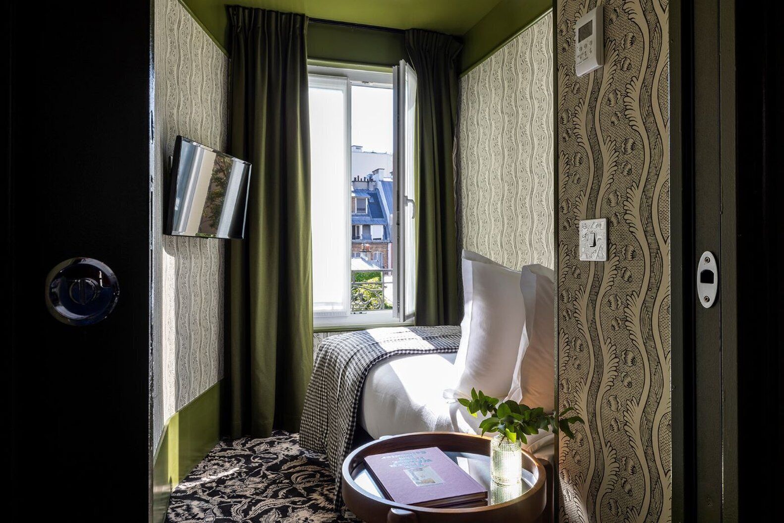 Hotelkamer met een bed, een nachtkastje met een boek en een vaas, een raam met gordijnen, en behang met een patroon.