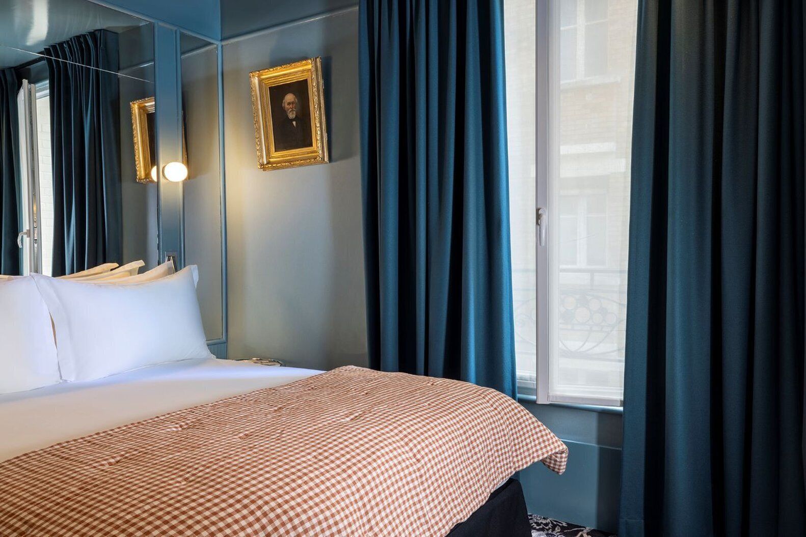 A hotel room with a large bed, a checkered throw blanket, dark curtains, and a framed picture on the wall.