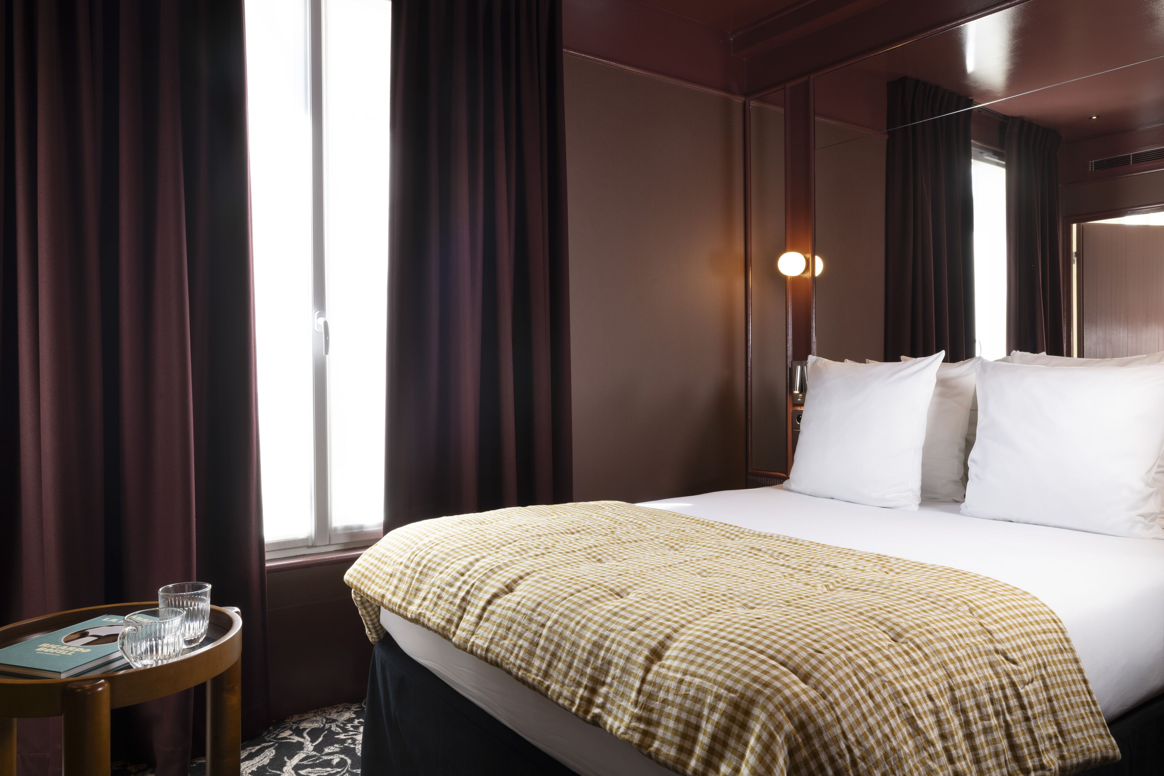 Habitación de hotel con una cama grande, una mesita de noche con un libro y gafas, y una ventana con cortinas.