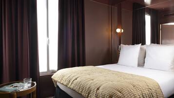 Habitación de hotel con una cama grande, una mesita de noche con un libro y gafas, y una ventana con cortinas.