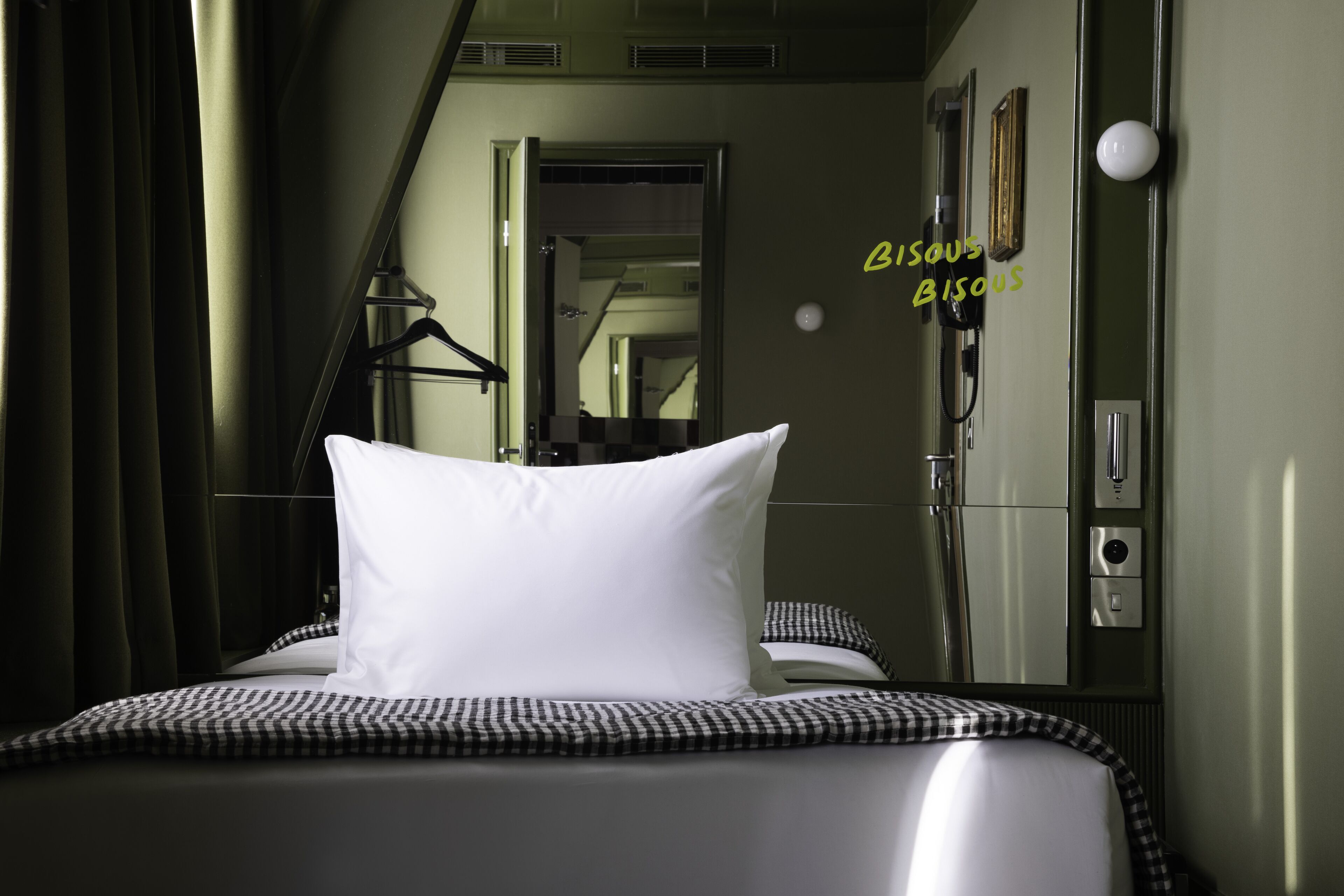 Una habitación de hotel con una cama, una colcha a cuadros, un almohadón blanco, una lámpara colgante, un espejo y una puerta que da a otra habitación.
