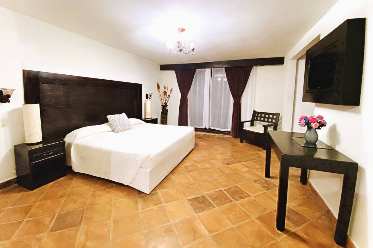 A hotel room with a large bed, a bedside table, a chair, a small table with a vase, a TV mounted on the wall, and a window with curtains.