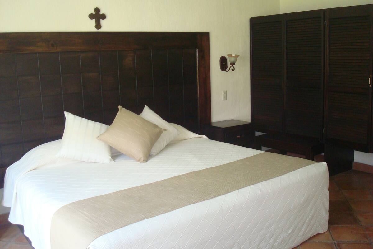 A bedroom with a large bed, a wooden headboard, a cross on the wall, and a nightstand with a lamp.