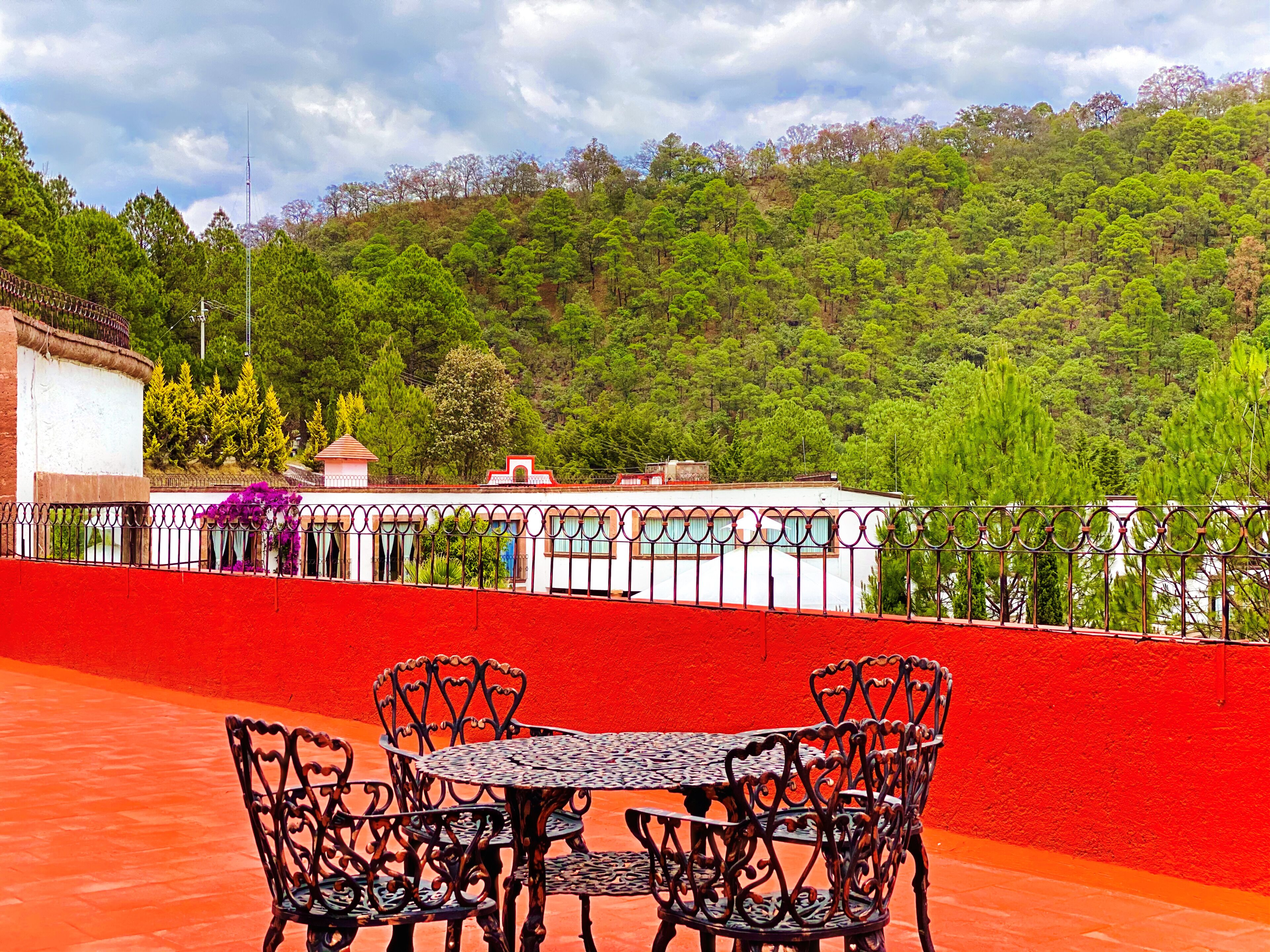Una terraza con una mesa y sillas, un muro rojo y vistas a una colina verde con árboles.