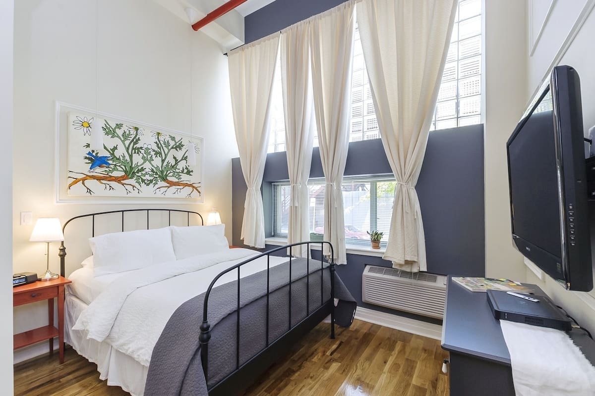 A bedroom with a bed, bedside table, lamp, artwork, windows with curtains, and a TV.