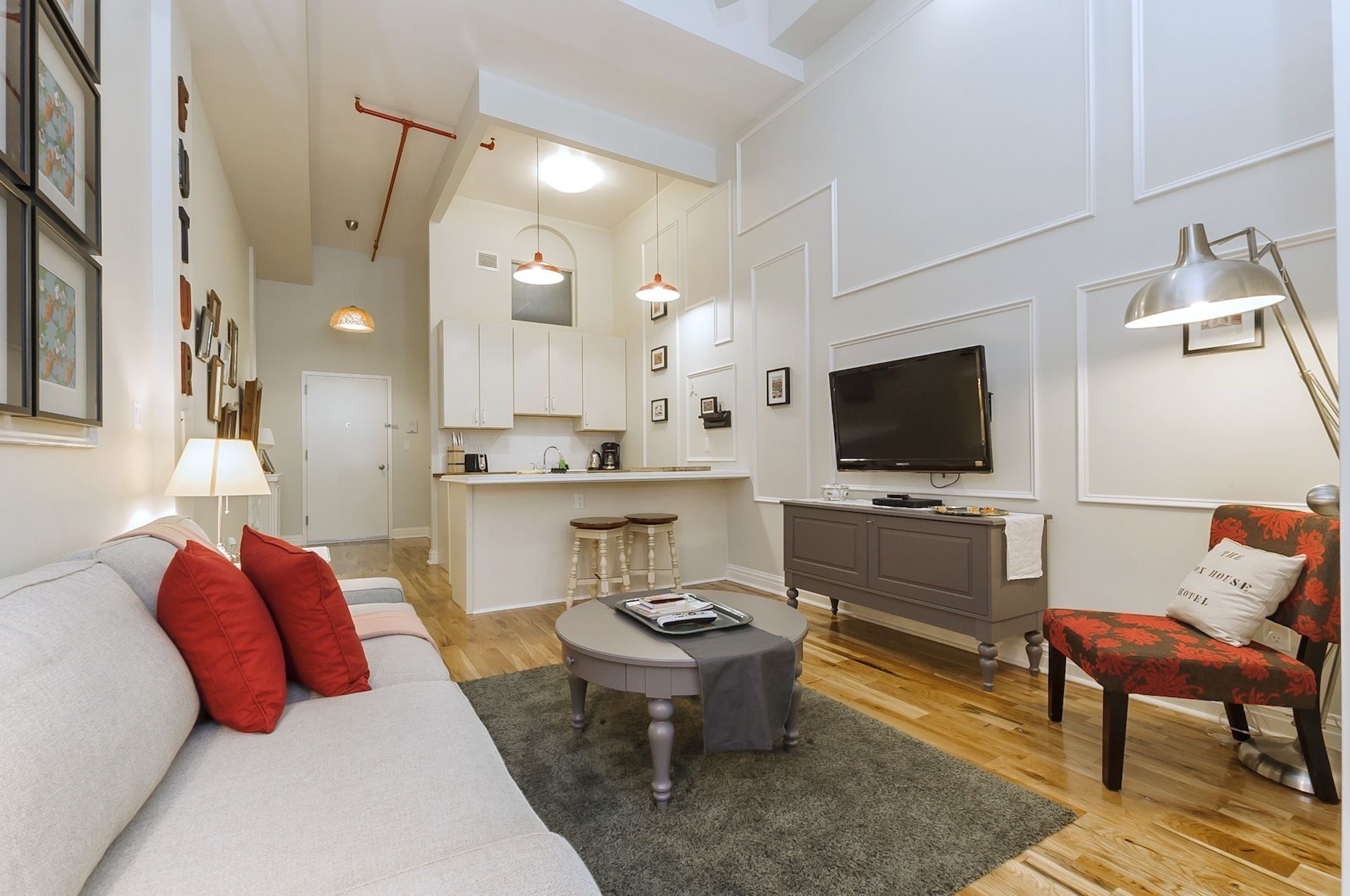 A modern living room with a sofa, red cushions, a coffee table, and a TV. There is a kitchen area with cabinets and a dining table.