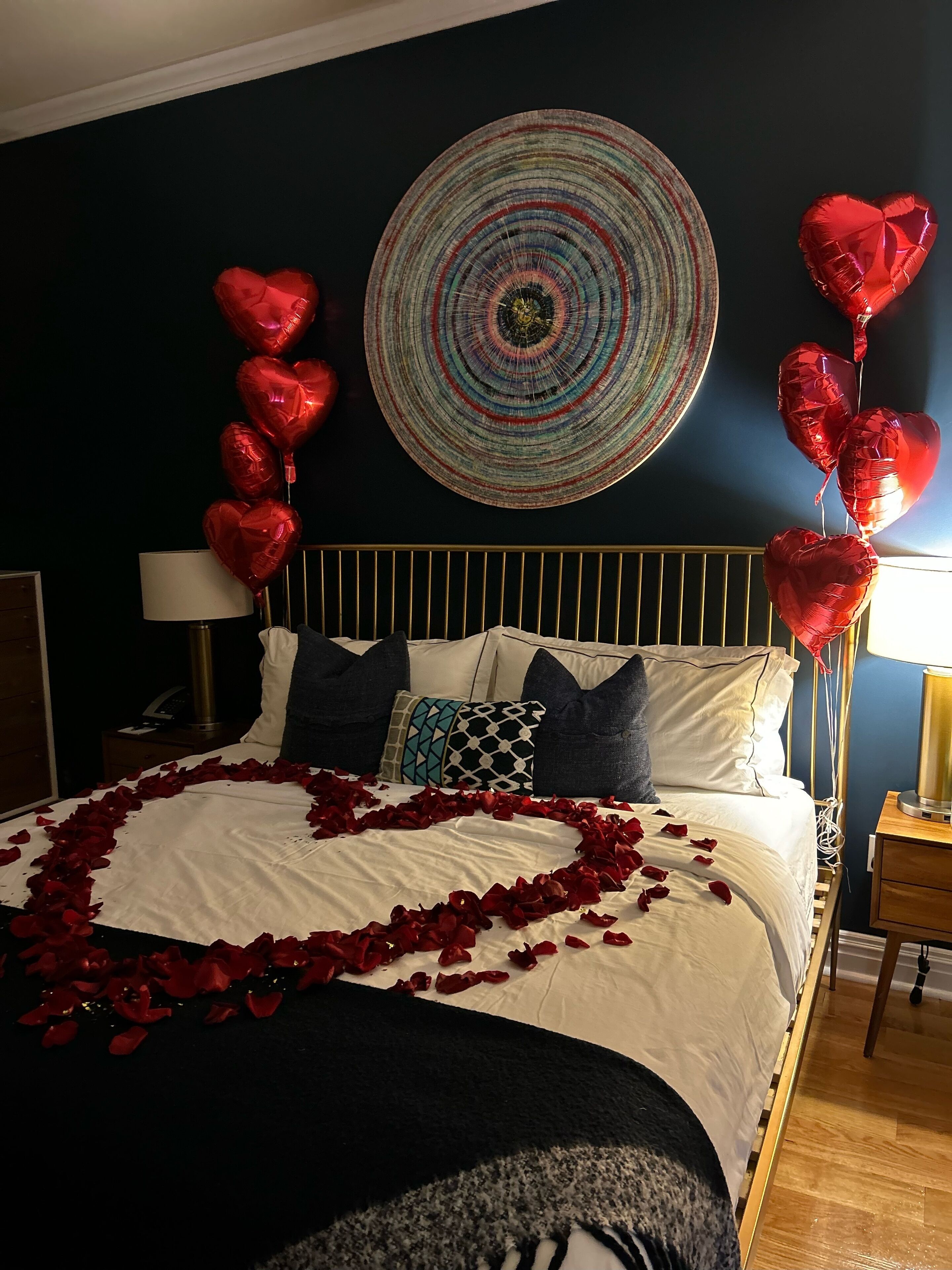 A bed with a heart-shaped arrangement of rose petals, red heart-shaped balloons, and a circular wall hanging.