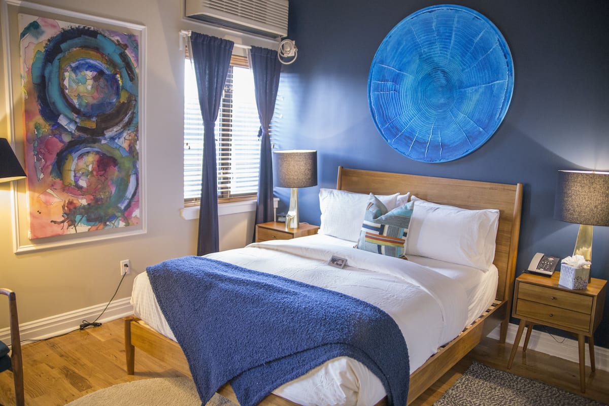 A neatly made bed with a blue blanket, a large abstract painting on the wall, and a round blue artwork above the bed.