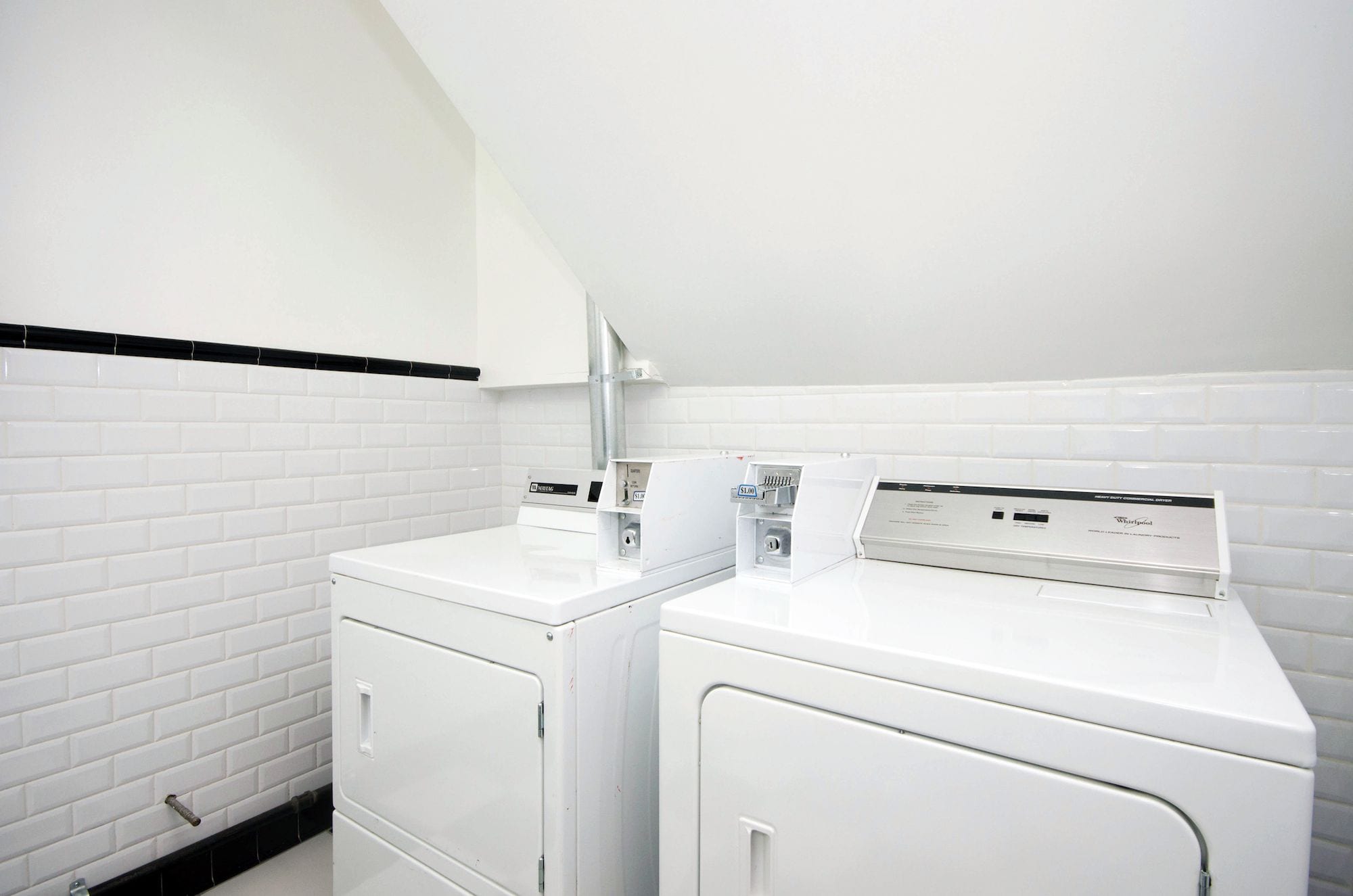 A laundry room with two stacked front-loading washers and dryers, white tiled walls, and a black baseboard.