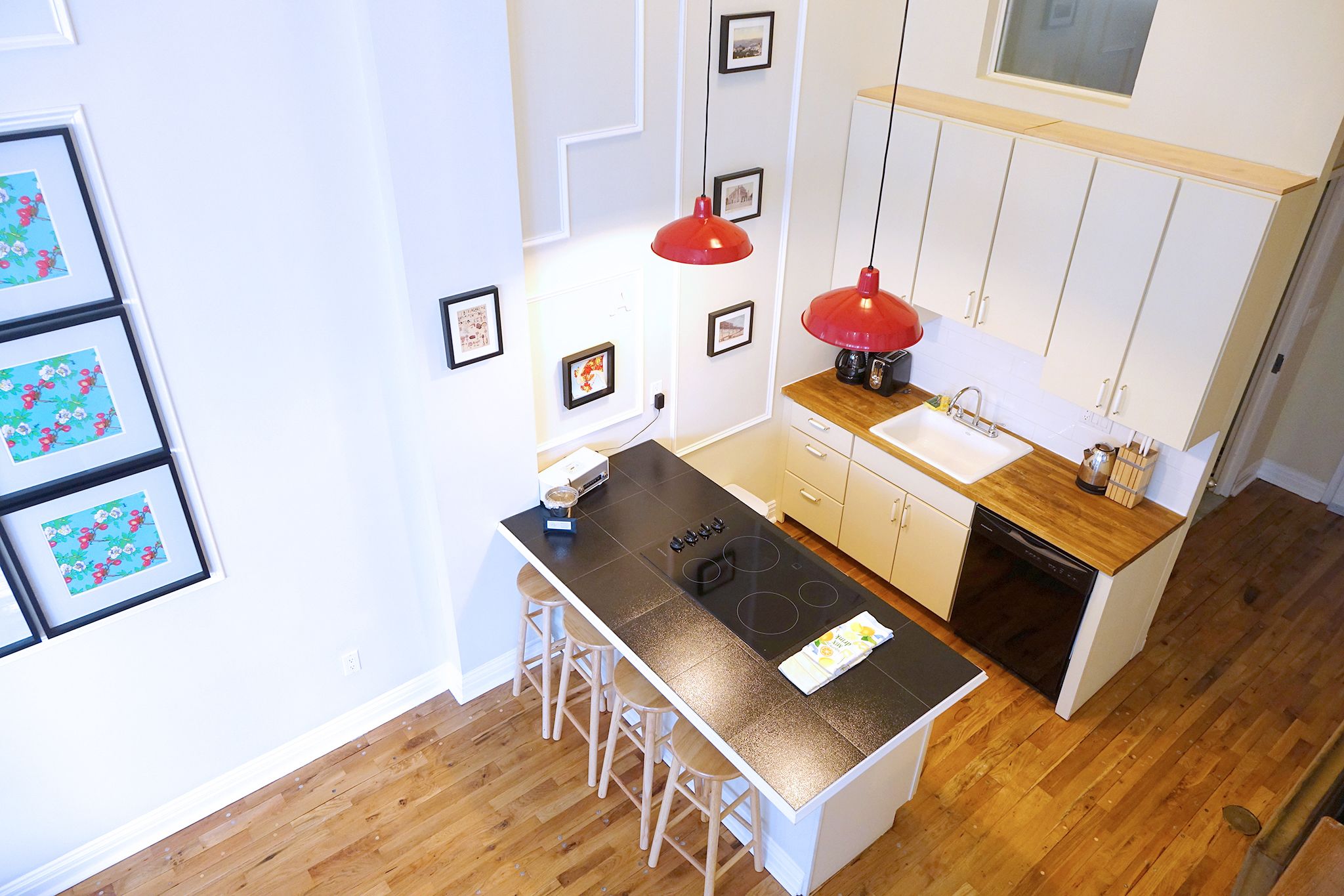 A compact kitchen with a central island, white cabinetry, a red pendant light, and framed pictures on the wall.