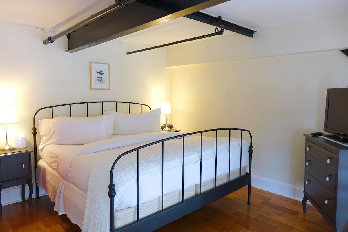 A bedroom with a metal-framed bed, a bedside table with a lamp, a dresser, and a framed picture on the wall.