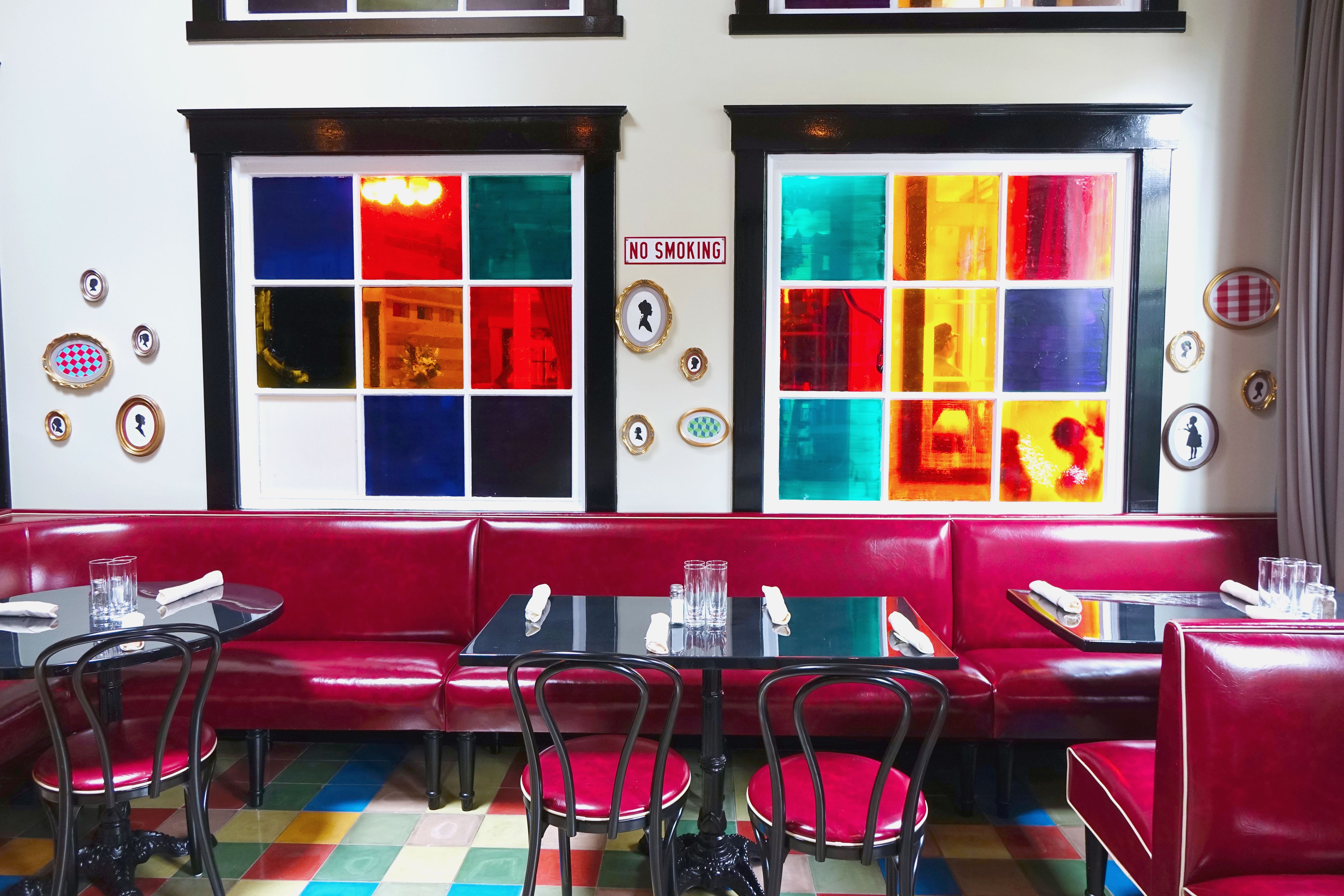Un restaurante con asientos de cuero rojo, mesas y sillas negras. Se observan vidrieras de colores y un cartel de 'No Smoking' en la pared.
