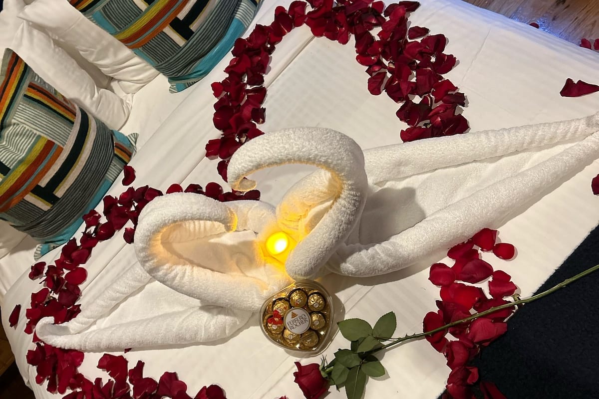 A bed with rose petals arranged in a heart shape, a bottle of Ferrero Rocher chocolates, and a lit candle.