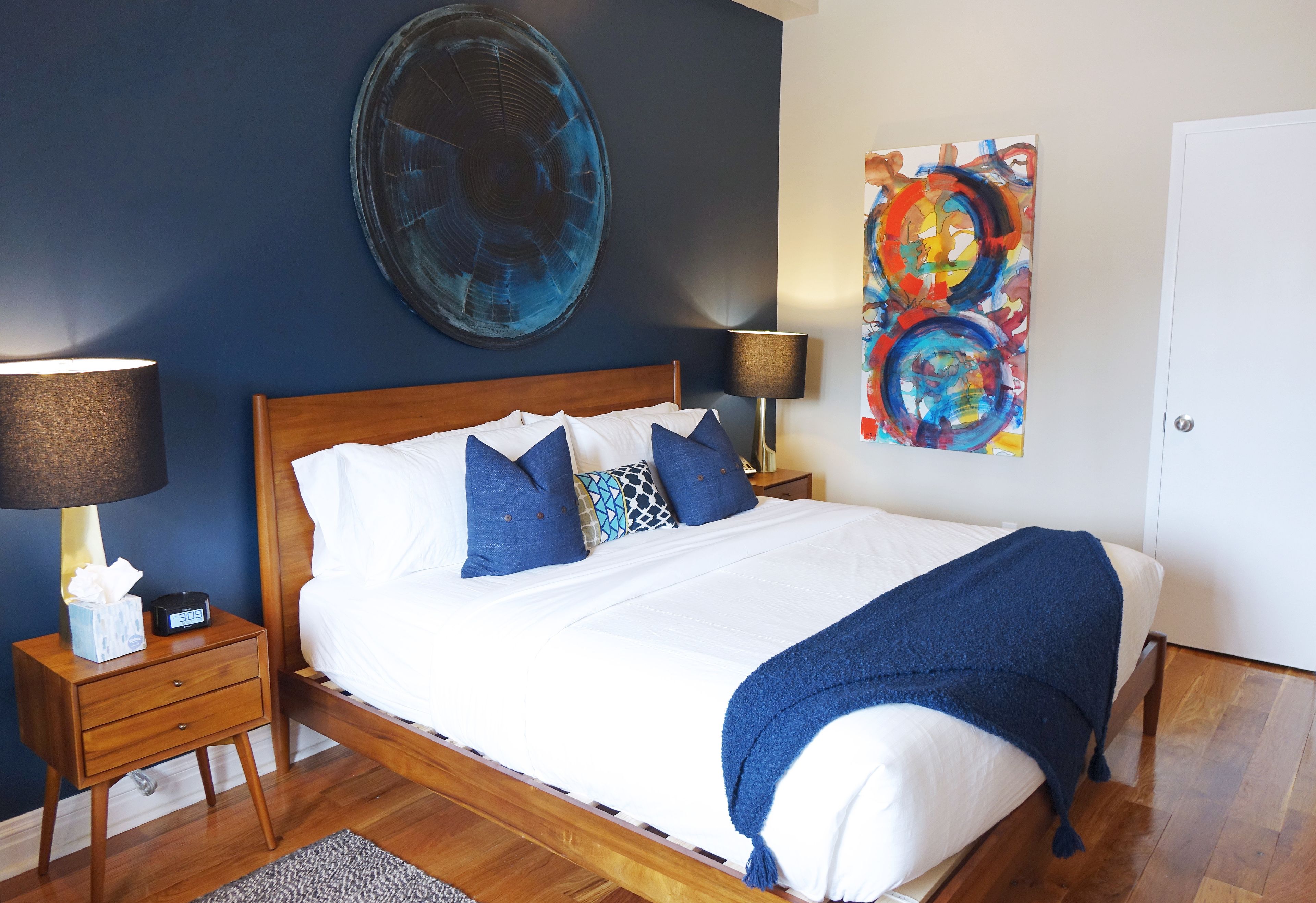 A neatly made bed with white and blue bedding, a blue throw blanket, and a wooden headboard. There are two bedside tables with lamps and a small bedside table with a tissue box and a clock.