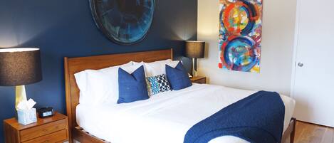 A neatly made bed with white and blue bedding, a blue throw blanket, and a wooden headboard. There are two bedside tables with lamps and a small bedside table with a tissue box and a clock.