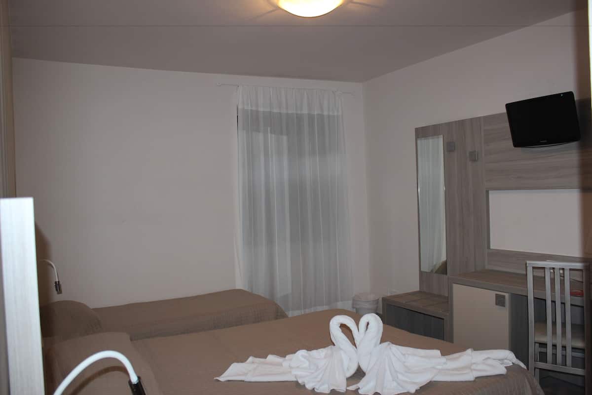 Habitación de hotel con dos camas, un televisor montado en la pared y una ventana con cortinas translúcidas.