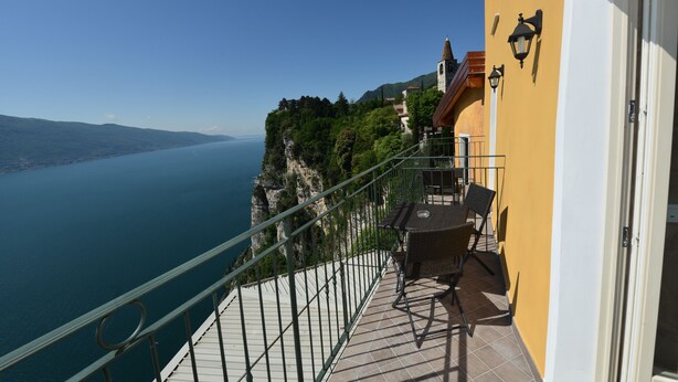 A balcony with a view of a lake and mountains, a table and chairs, and a building with a yellow wall and a door. - Miralago (Tremosine)