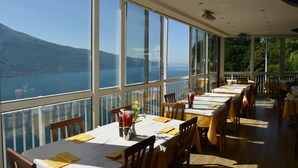 A restaurant with large windows offering a view of a lake and mountains. Tables are set with yellow napkins and menus. - Miralago (Tremosine)