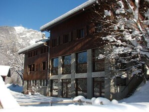 Un bâtiment moderne doté de grandes fenêtres et d’une façade en bois, entouré d’arbres et de montagnes couverts de neige.