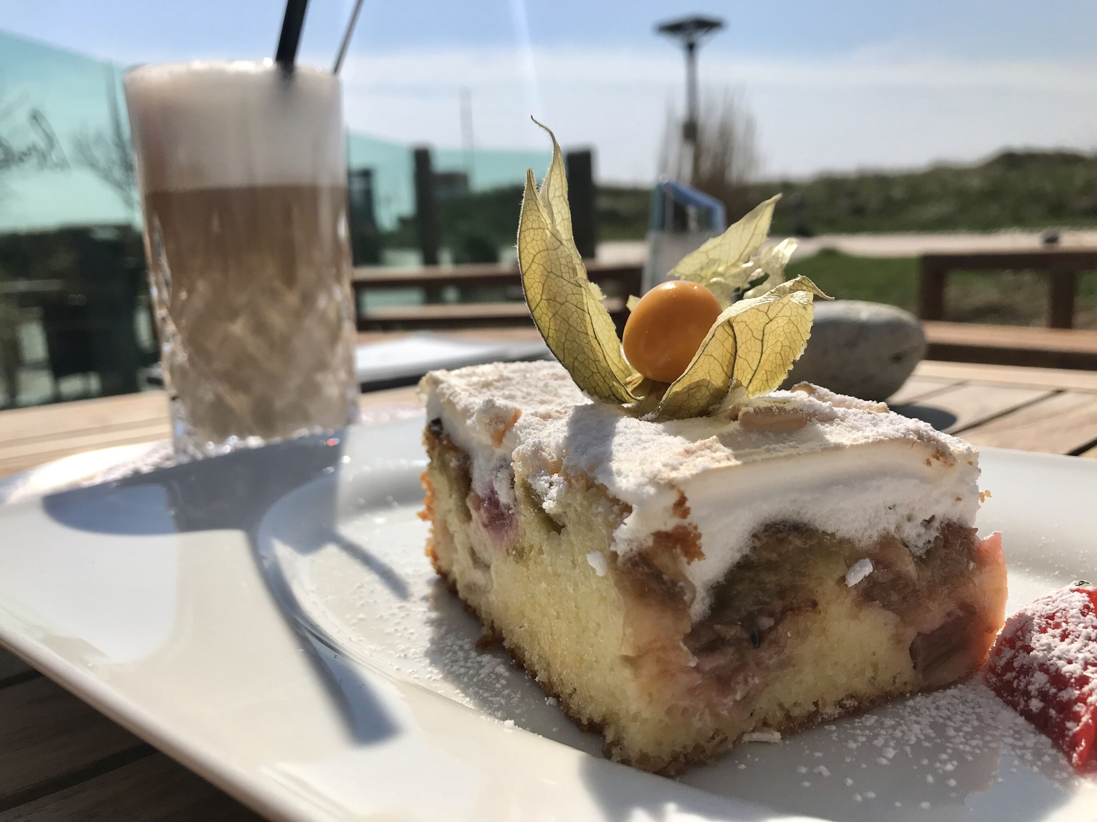 A slice of cake with powdered sugar and a fruit garnish on a white plate, accompanied by a glass of a beverage with a straw.