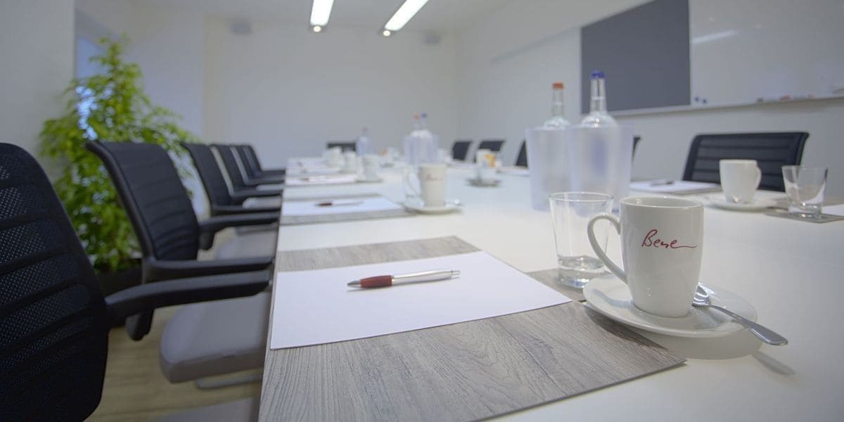 Un salón de conferencias con una larga mesa, sillas y una taza de café con la palabra "Bene".