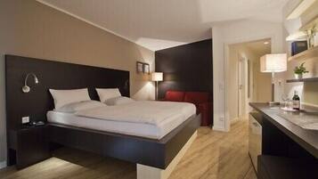Une chambre d’hôtel moderne dotée d’un grand lit, d’une table de chevet, d’un bureau avec une chaise et d’un petit coin salon avec un fauteuil rouge.