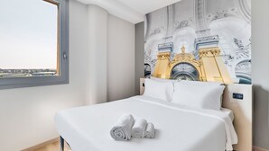 Una habitación de hotel moderna con una cama grande, vistas de la ciudad a través de una ventana y un mural con detalles arquitectónicos en la pared.