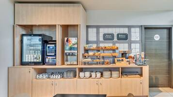 Un área de desayuno con una máquina de café de autoservicio, un refrigerador con productos y una variedad de pasteles y productos horneados.