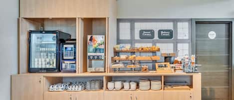 A breakfast area with a self-service coffee machine, a refrigerated display case, and a variety of pastries and baked goods.