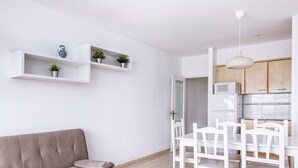 A modern living room with a sofa, a dining table with chairs, white shelves with plants, and a kitchen area with cabinets and a microwave. -  RVApartaments Club Torrevella (Torroella de Montgrí)