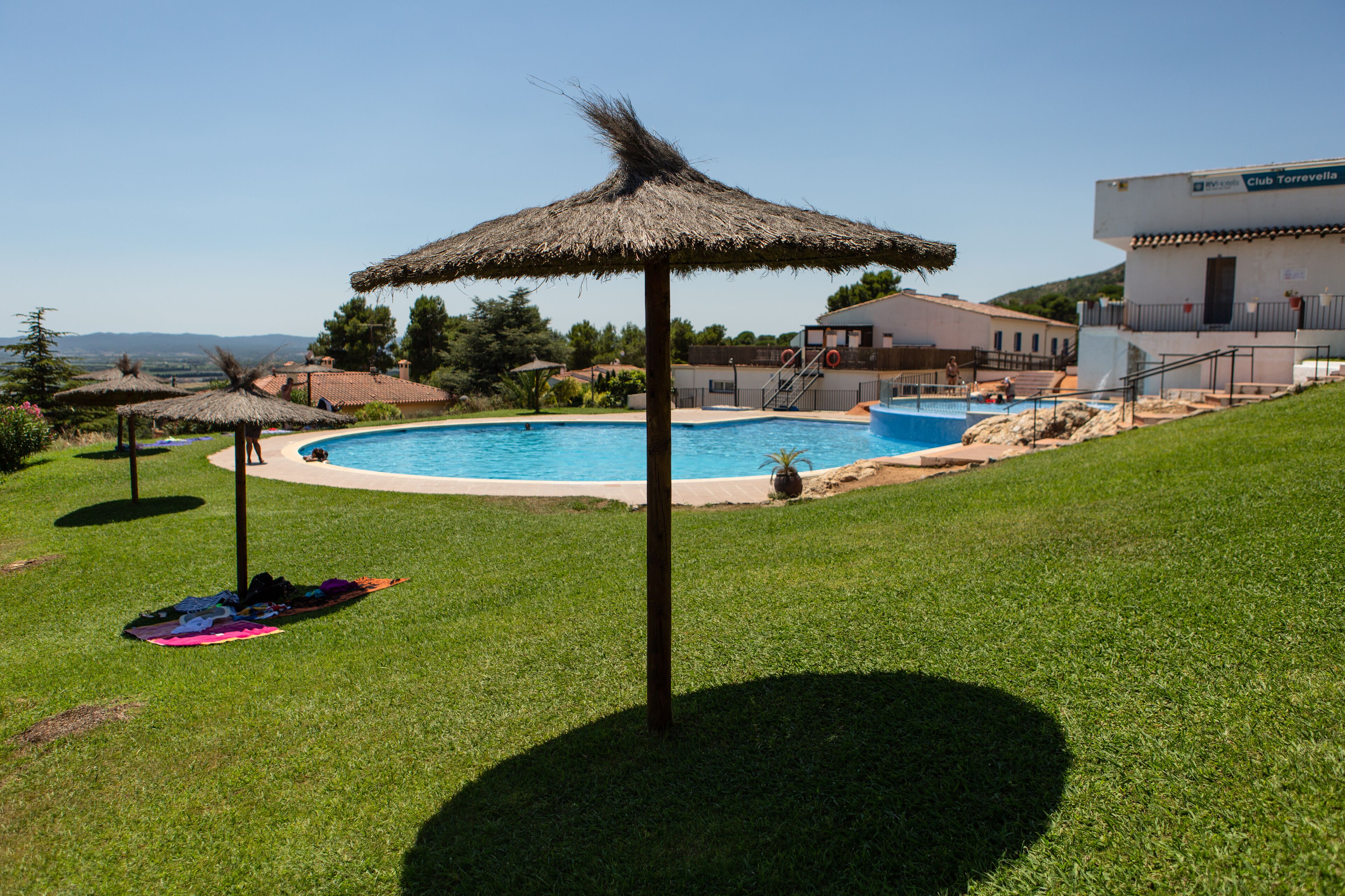 Ein Swimmingpool mit einem Strohdachschirm, einer Rasenfläche und im Hintergrund ein Gebäude mit der Aufschrift 'Club Torrevella'.